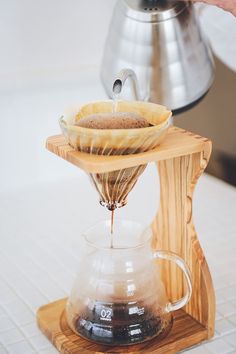 a person pours coffee into a glass cup on a wooden stand with a metal kettle in the background