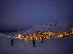 two people walking up the side of a snow covered mountain at night with city lights in the distance