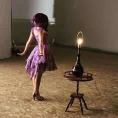 a woman in a purple dress standing next to a lamp on a tripod stand