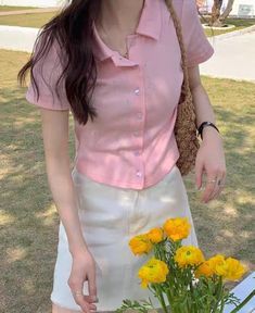 a woman in pink shirt and white skirt standing next to yellow flowers on table outdoors