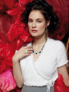 a woman in white shirt and grey skirt posing with red flowers behind her, wearing jewelry