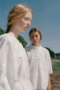 two women standing next to each other in front of trees and dirt ground, one wearing a white blouse