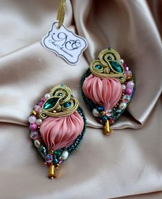 two pink and green earrings on top of a white satin bag with a tag hanging from it