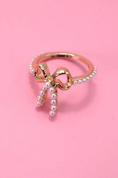 Introducing our Gold Pearl Bow Ribbon Ring, a delightful fusion of elegance and charm. This exquisite ring features a lustrous pearl nestled within a gracefully crafted bow and ribbon design, adding a touch of sophistication to your jewelry collection. Elevate your style with the delicate beauty of the Gold Pearl Bow Ribbon Ring, a symbol of refined taste and grace. DIMENSION: size: 7 Metal finish: Gold Plating product: Lead & Nickle Compliant anti-tarnish: Double E-coating Elegant White Jewelry With Pink Bow, Gold Pearl Rings For Party, Party Gold Rings With Pearl, Party Gold Pearl Rings, Elegant Adjustable Jewelry With Pink Bow, Elegant Adjustable Jewelry With Satin Bow, Party Pearl Jewelry With Bow, Wedding Jewelry With Pink Bow, Elegant Gold Jewelry With Pink Bow