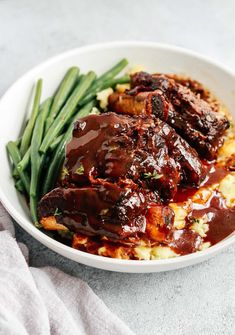 a white bowl filled with meat and potatoes covered in gravy next to green beans