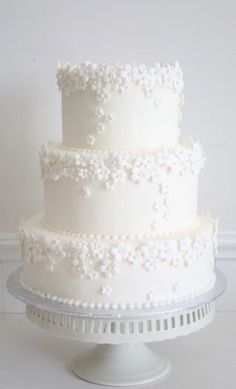 a three tiered white wedding cake sitting on top of a glass platter in front of a wall