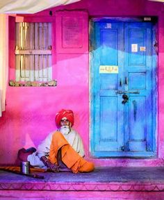 a man sitting on the steps in front of a building with blue doors and pink walls