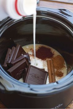 someone pouring milk into a slow cooker filled with chocolate and cinnamon sticks, next to some cocoa bars