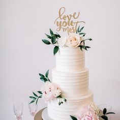 a white wedding cake with flowers and greenery on the top is decorated with gold lettering love you most