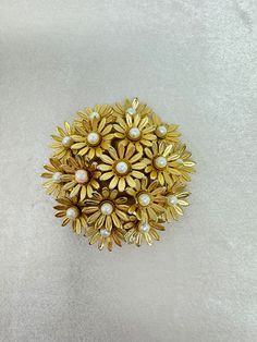 "Add a touch of vintage charm to your collection with this stunning Daisy Flower Brooch. Crafted in a beautiful gold tone, this brooch features a captivating daisy design with a faux pearl accent.  Measuring at 2 1/4\" in diameter and 3/4\" in height, this brooch is the perfect size to adorn your favorite outfits, bags, or hats.  Please note that this brooch is unsigned, adding to its unique appeal and vintage allure.  Don't miss out on this exquisite piece that will surely make a statement wherever you go. Order now and add a touch of timeless elegance to your style. Enjoy the convenience of free shipping within the United States on all of my exquisite jewelry pieces. With priority mail, your order will be swiftly delivered to your doorstep.  Take a moment to explore my other captivating Vintage Gold Flower Shaped Brooch, Gold Flower-shaped Brooch With Floral Decoration, Vintage Yellow Gold Flower Brooch, Vintage Yellow Gold Flower Brooches, Vintage Gold Flower Brooch, Vintage Gold Flower Brooches, Daisy Design, Exquisite Jewelry, Bridesmaids Gifts