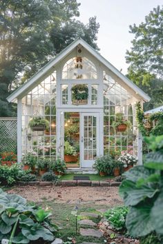 a white greenhouse with potted plants in the front and windows on each side, surrounded by greenery