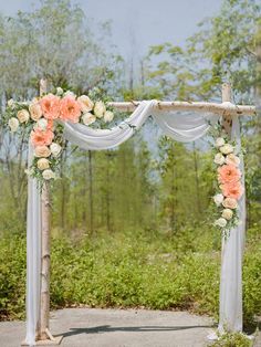 an outdoor wedding ceremony with white and peach flowers on the arch, greenery and trees in the background