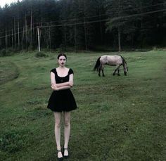 a woman in a black dress standing next to a horse on a lush green field