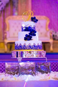 a three tiered wedding cake with blue flowers on the top and purple ribbon at the bottom