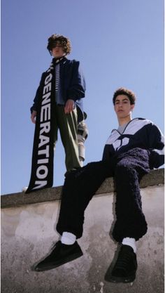 two young men sitting on top of a cement wall next to each other wearing sweaters and pants