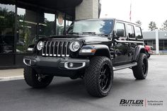 a black jeep is parked in front of a building with large tires on it's rims