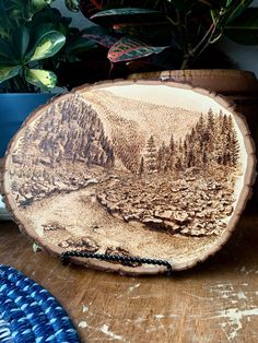 a wooden plate with a landscape painted on it sitting on a table next to potted plants