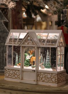 a glass house with deer and christmas trees in the front, on a wooden table