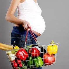 a pregnant woman holding a shopping basket full of fruits and vegetables in her belly, against a gray background