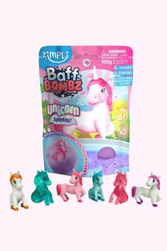 three small toy unicorns next to a bag of bath bombz on a pink background