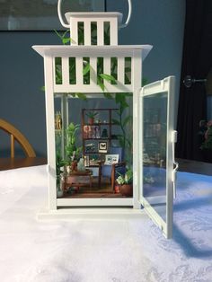 a miniature house with plants in it on a white tableclothed surface, next to a chair and window