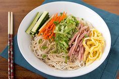 a white bowl filled with noodles and veggies next to chopsticks on a blue napkin