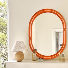 an orange round mirror sitting on top of a white mantle next to a lamp and table