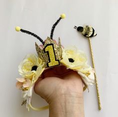 a hand holding a yellow and black number one crown with flowers on it, next to a wand