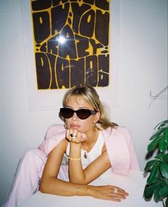 a woman in sunglasses sitting on a bed with her hand under her chin and looking at the camera