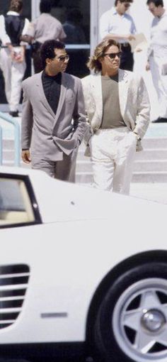 two men walking next to a white sports car in front of some people on the street