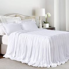 a white bed with ruffled bedspread and pillows in a bedroom next to a window