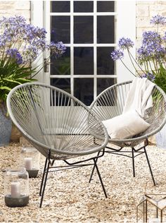 two chairs sitting on top of a gravel covered ground next to flowers and candles in front of a window