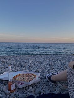 a person sitting on the beach with a pizza and soda in front of them at sunset