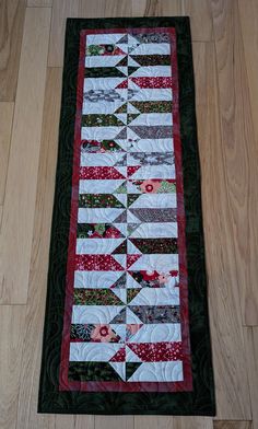 a quilted table runner with red and green flowers on the border, sitting on a wood floor