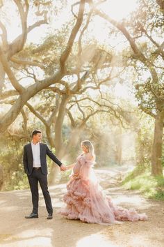 a bride and groom holding hands while walking down a dirt road in front of trees