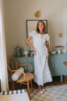 a woman in a white dress standing next to a dresser and chair with her hands on her hips