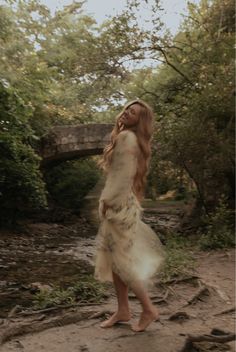 a woman in a white dress walking across a dirt road next to a river and bridge