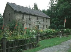 an old wooden house surrounded by lush green trees and flowers in front of a fence