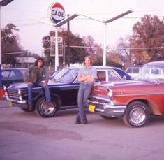 two men sitting on the hood of an old car in a gas station parking lot