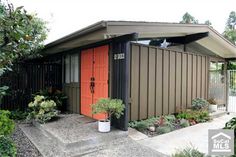 an orange door sits in front of a brown house with green trees and bushes around it