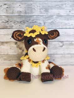 a crocheted cow with a yellow flower on its head sitting in front of a wooden wall