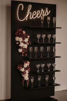 a black shelf with wine glasses and flowers on it next to a neon sign that says cheers