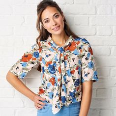 a woman standing in front of a white brick wall wearing a floral blouse and jeans