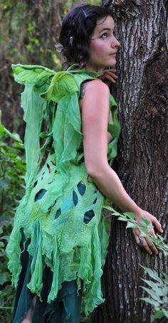 a woman in a green dress standing next to a tree with her hands on the trunk