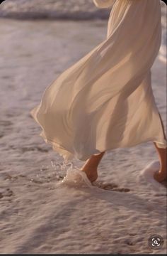 a woman in a white dress walking on the beach with her feet in the sand