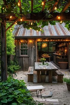 an outdoor kitchen with lights hanging from the roof and wooden benches underneath it, surrounded by greenery