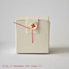 a small white box with a red ribbon and button on the side that says by / between the lines /