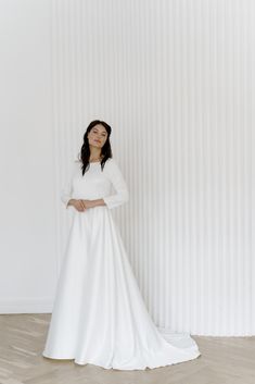 a woman standing in front of a white wall wearing a long sleeved wedding dress