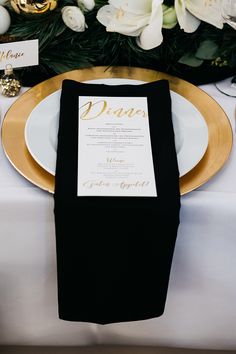 a place setting with black napkins and white flowers on the table, along with gold chargers