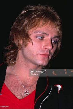 a young man with long hair wearing a red shirt and black jacket looking at the camera